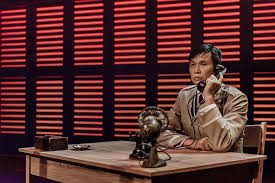 Playing a police officer named Tommy Lan, John Ng sits at a desk in the Kowloon police station. 